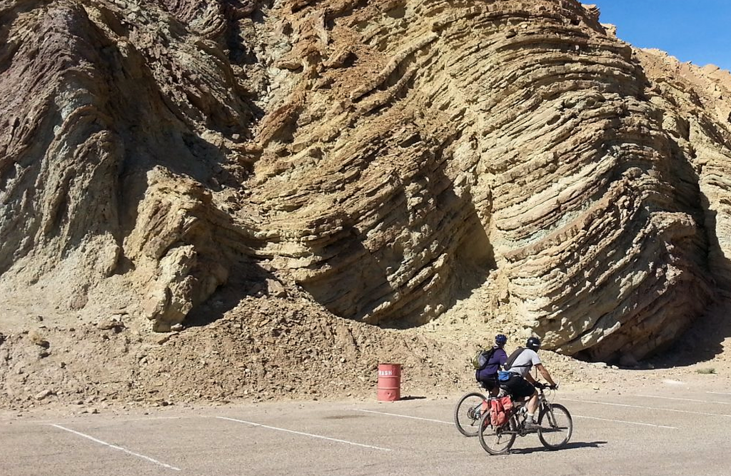 Two bicycle riders riding by a big Calico rock in a parking lot.