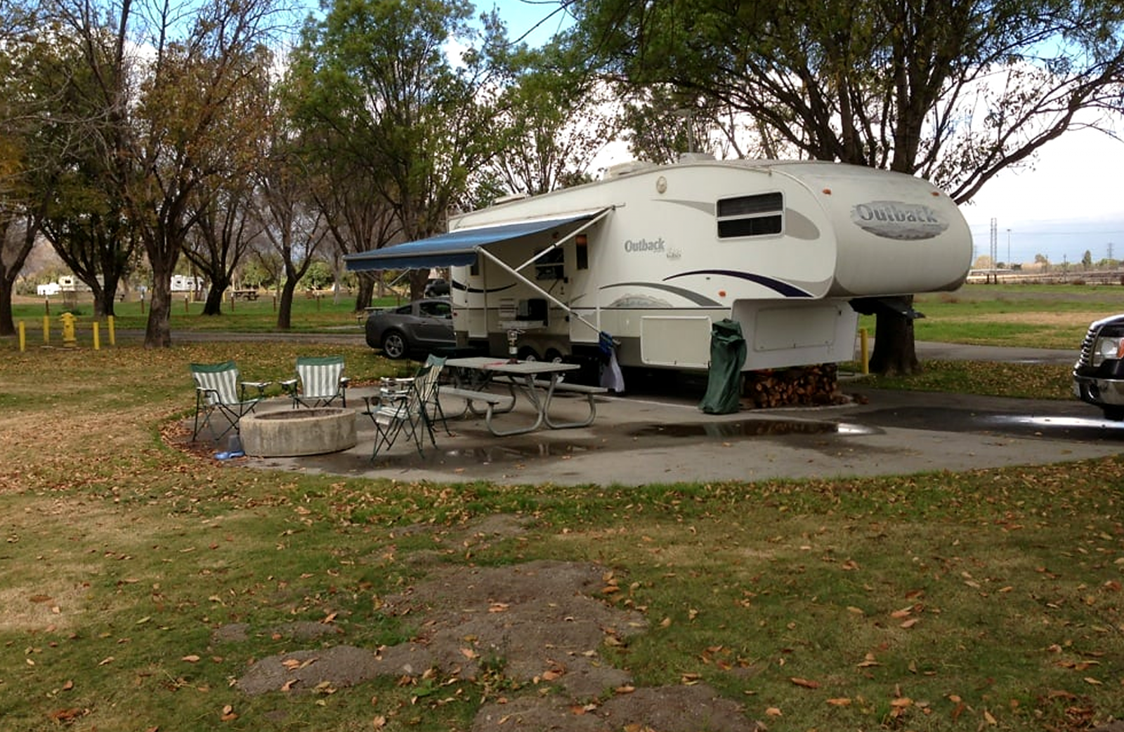 A single camping trailer sits on a concrete slab near grass and a fire ring at Prado park.