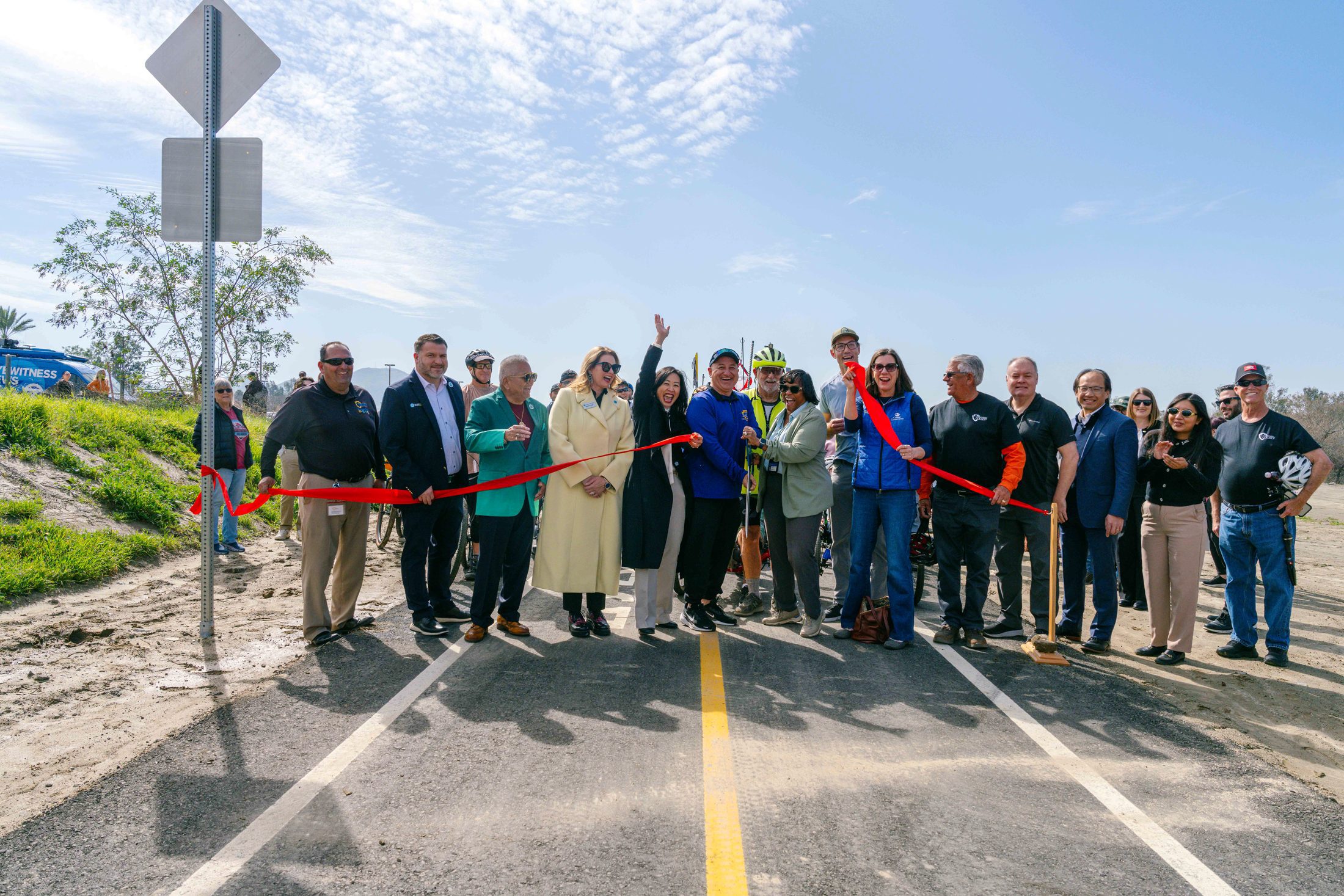 A group of about 20 people cheer and smile while standing on a portion of the Santa Ana River Trail bike path behind a large red ribbon that is cut into two pieces.