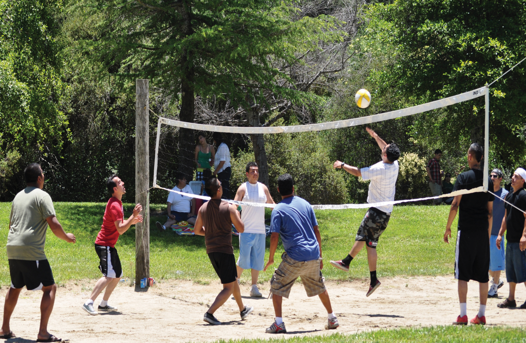 A group of young people playing outdoor volleyball on a sand court surrounded by grass.