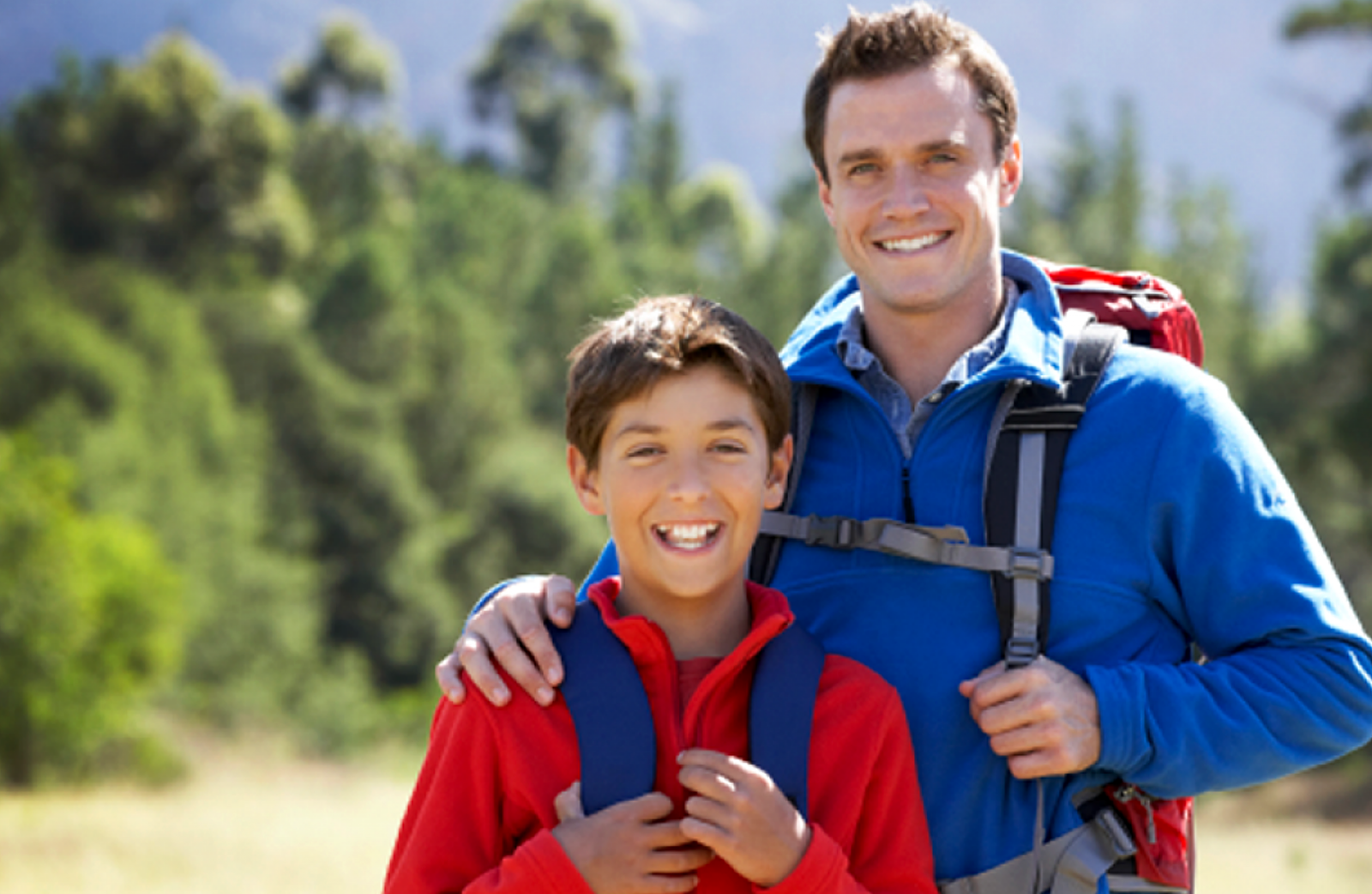 A father with his arm on his son's shoulder and son smile in a park setting.