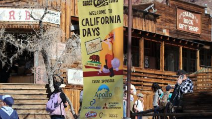 A wide photo of the California Days banner hanging in the middle of Calico Ghost Town with people walking by in front of the R&D Fossil Shop.