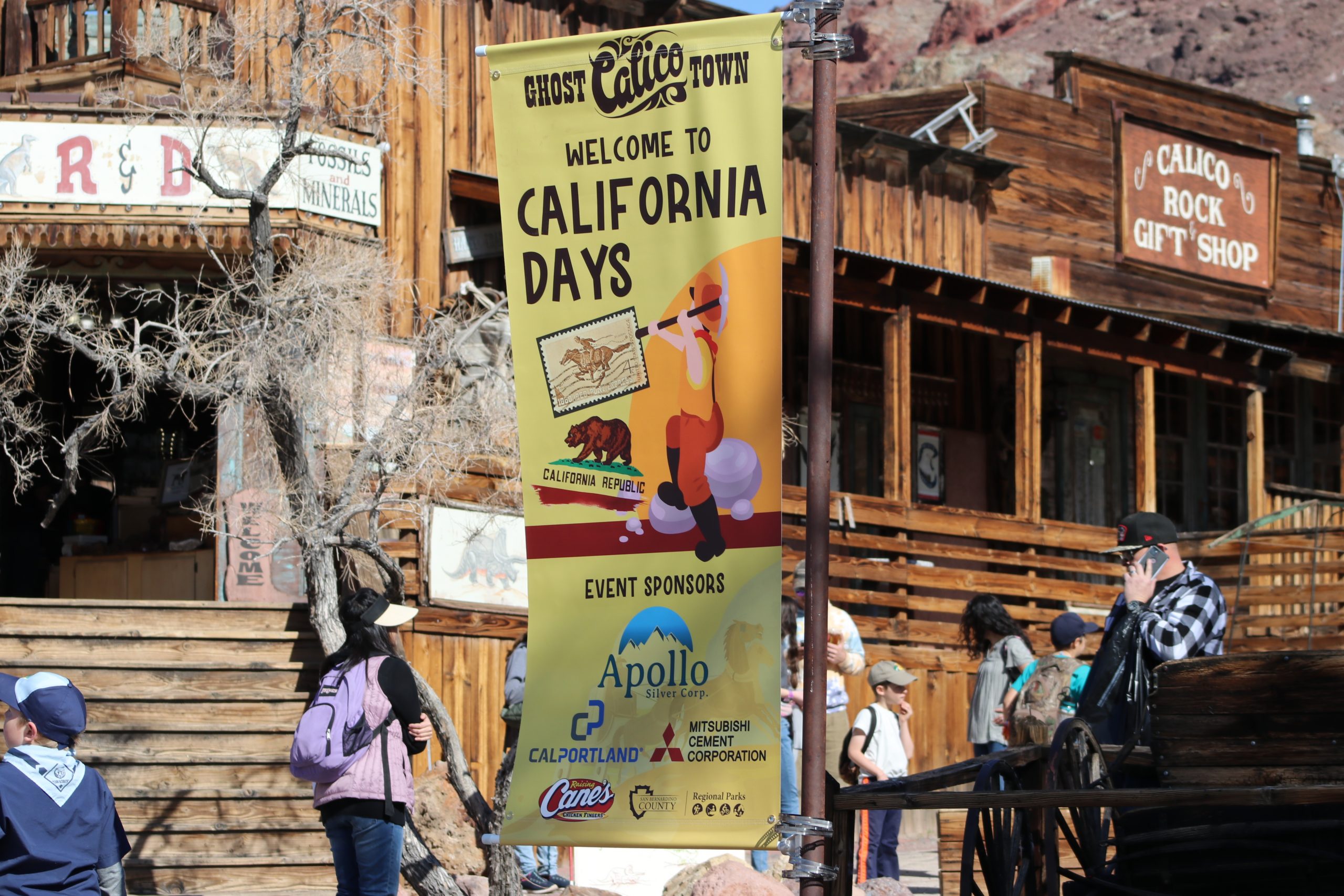 A wide photo of the California Days banner hanging in the middle of Calico Ghost Town with people walking by in front of the R&D Fossil Shop.