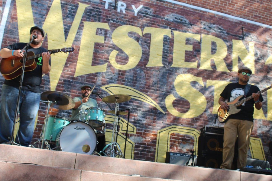 Three male musicians perform onstage in front of a brick building wall that says Western Store in vintage yellow lettering.
