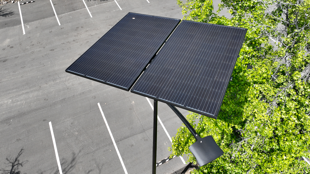 A photo of a new black solar power light pole flat panel with a LED light extending downward taken overhead with the view of a parking lot beneath it and a tree on the right.