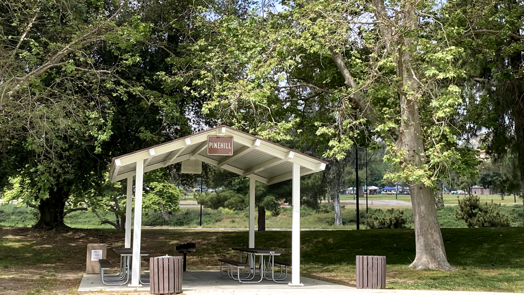 A photo of a small new picnic shelter with grass, trees and trashcans flanking the  shelter that says Pinehill.