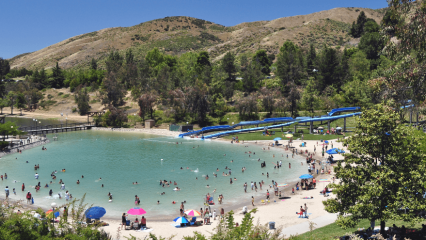 A overhead view of the pool at Yucaipa Regional Park with mountains and waterslides in the background with a blue sky. People are seen in the lagoon-style pool with people on the sandy beach area around the pool.