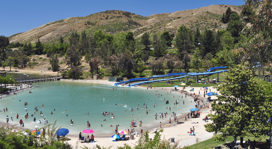A overhead view of the pool at Yucaipa Regional Park with mountains and waterslides in the background with a blue sky. People are seen in the lagoon-style pool with people on the sandy beach area around the pool.