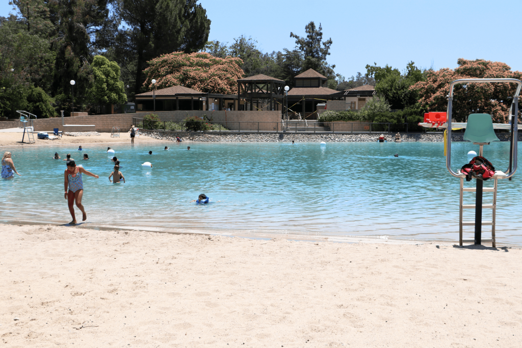 A photo of a little girl in her bathing suit coming out of the lagoon-style pool with sand in the foreground, blue sky and trees in the background with blue pool water.
