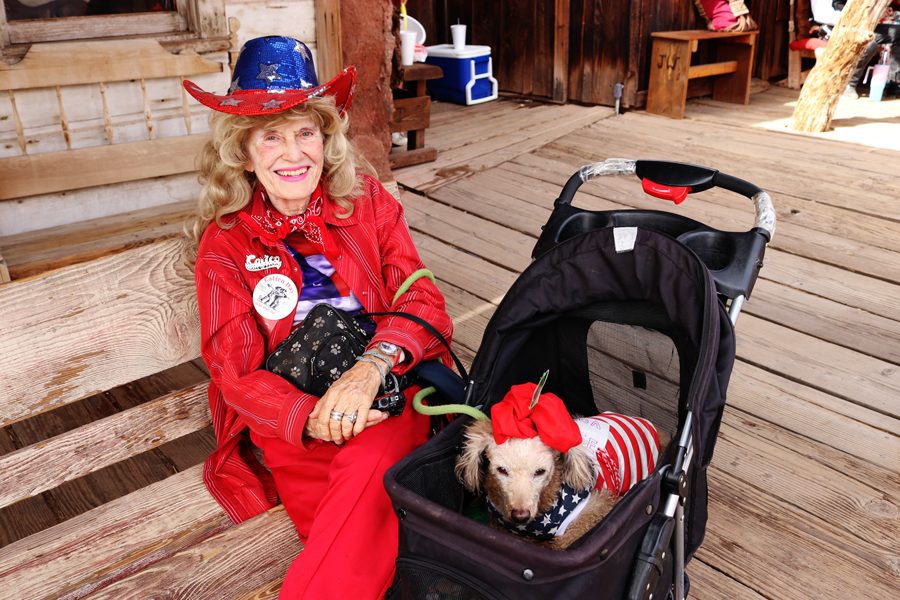 A woman dressed in red and blue costume with western hat and a poodle dress in same costume sitting in a stroller.