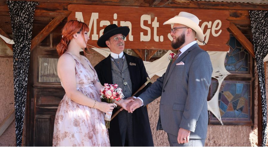 A woman and man stand facing each other on the Main Street Stage at Calico Ghost Town with a man dressed in 1800 period clothing standing in between looking at the man.