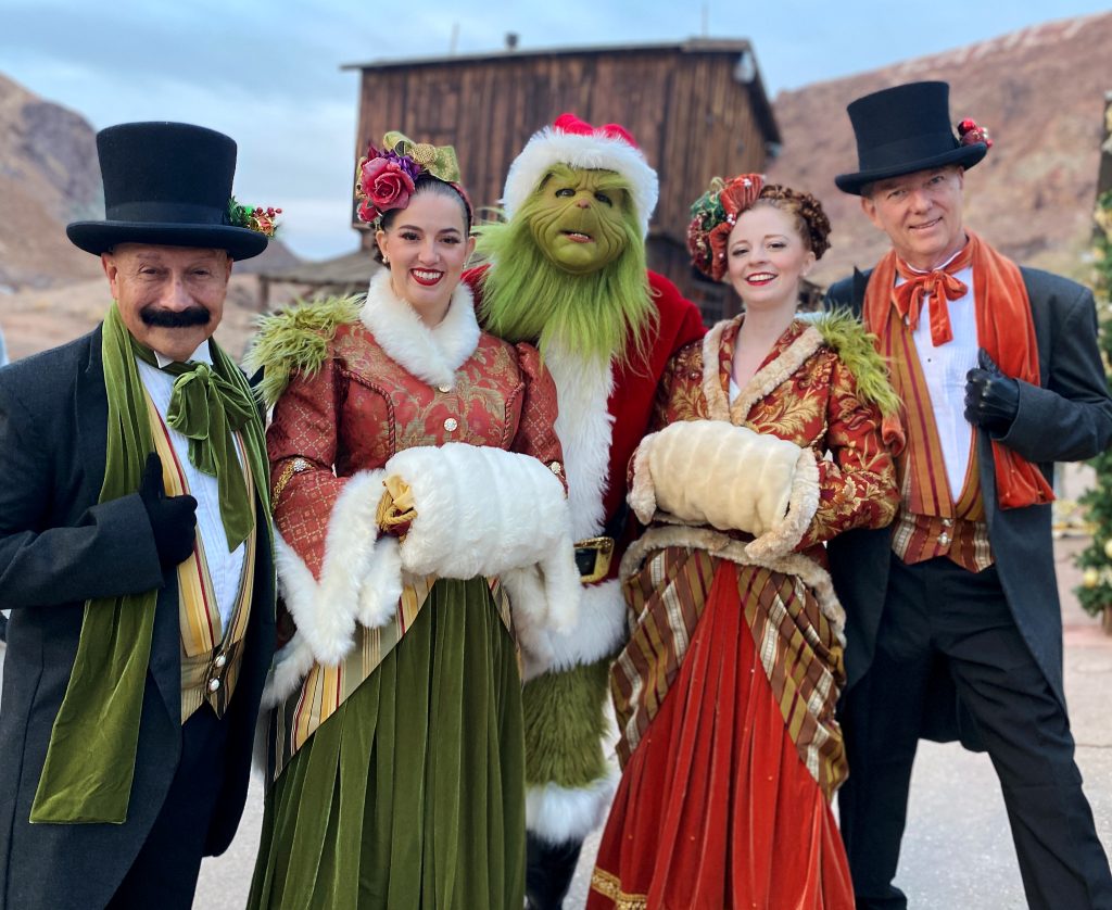 Four people dressed in 1800s period clothing stand with a person dressed as the Grinch in a Santa costume at Calico Ghost Town.