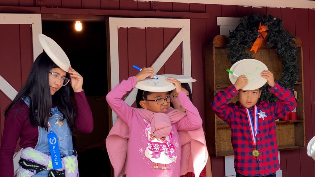 Three young girls hold paper plates on their head trying to draw with a pen at Calico Holiday Fest.
