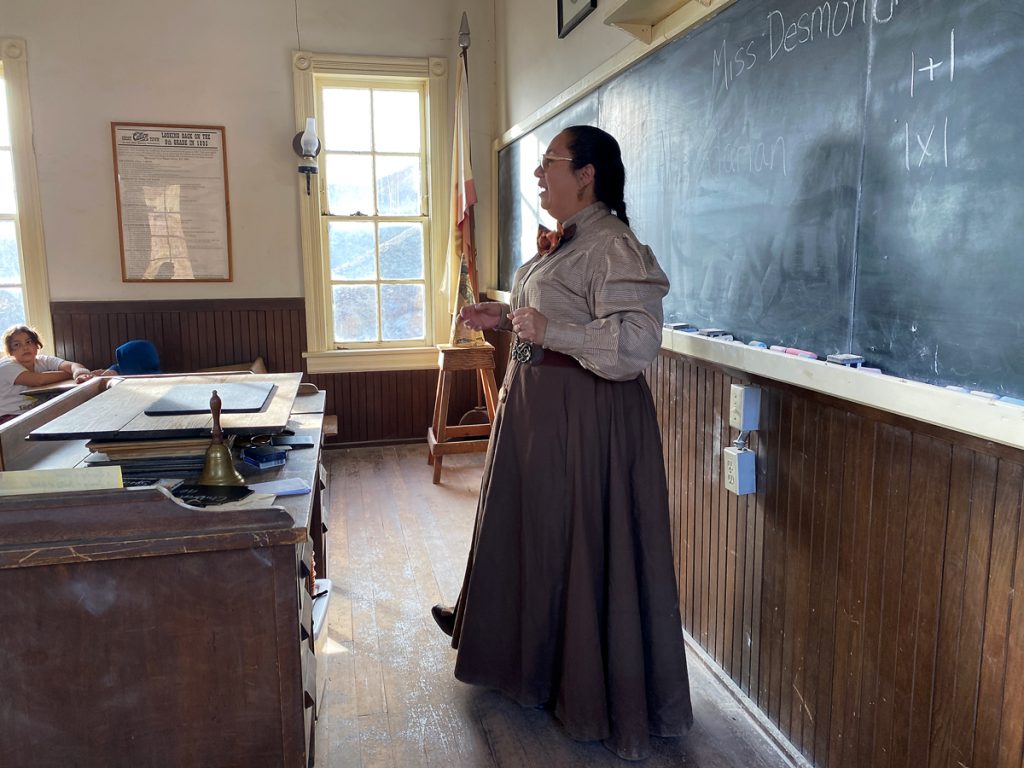 A woman stands in an old-fashioned schoolhouse wearing 1800s period clothing.
