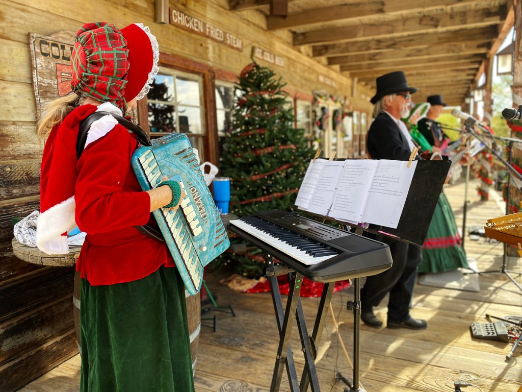 A woman dressed in holiday 1800s clothing hold an accordion on the porch with a man playing an instrument in the distance on the Calico House Restaurant at the Calico Holiday Fest.