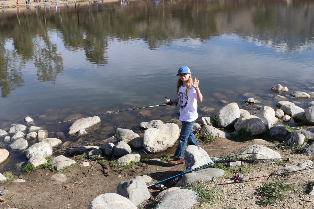 A girl waving near a lake while holding a fishing pole at Yucaipa Regional Park.