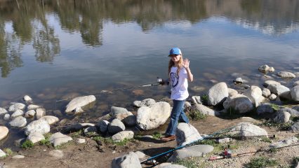 A girl waving near a lake while holding a fishing pole at Yucaipa Regional Park.