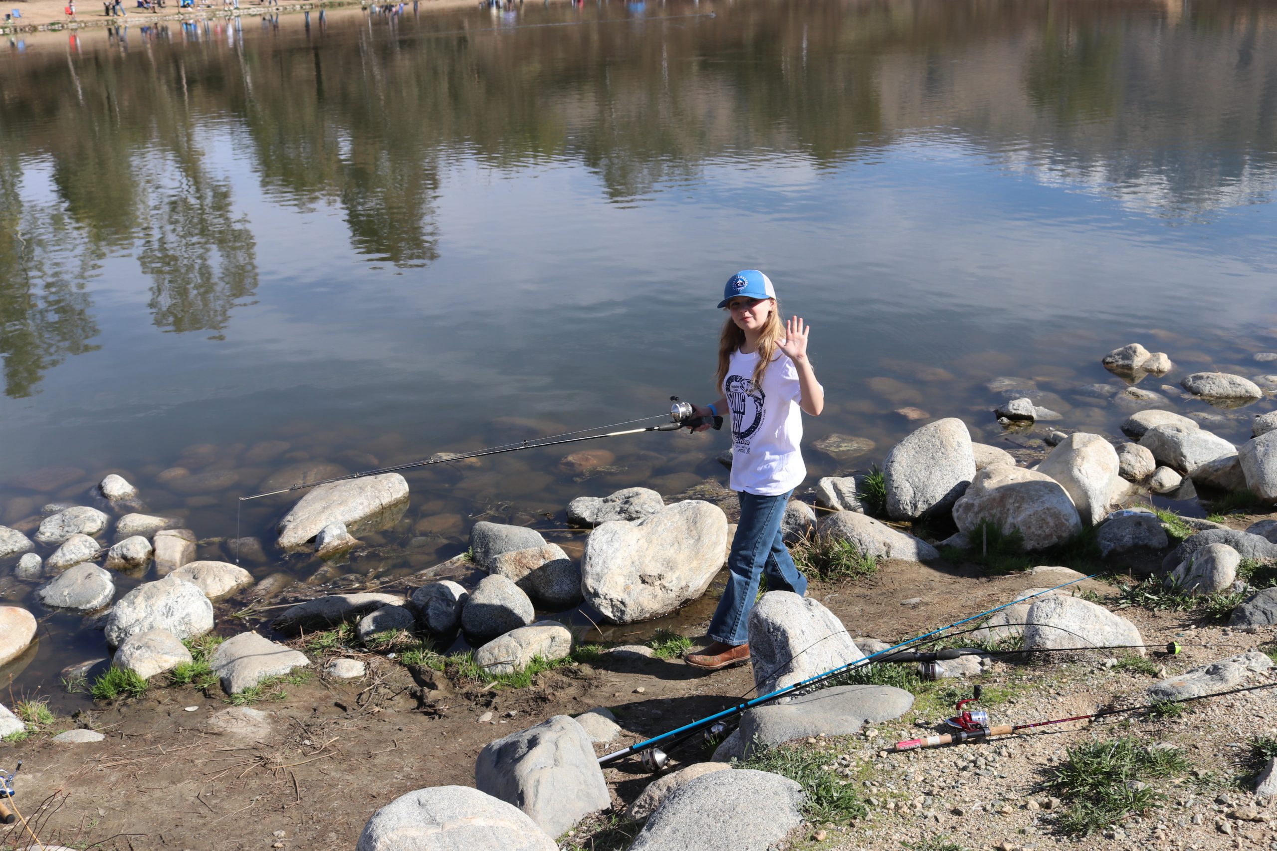 A girl waving near a lake while holding a fishing pole at Yucaipa Regional Park.