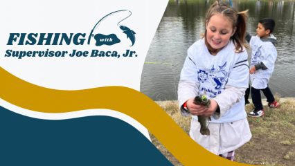 A young girl smiles while holding a fish and a young boy holds a fishing pole behind her near a lake. The word Fishing with Supervisor Joe Baca, Jr. is on the left.
