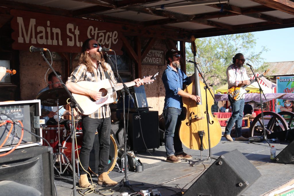 Three male musicians play instruments on a stage while another male drummer plays a drum set.