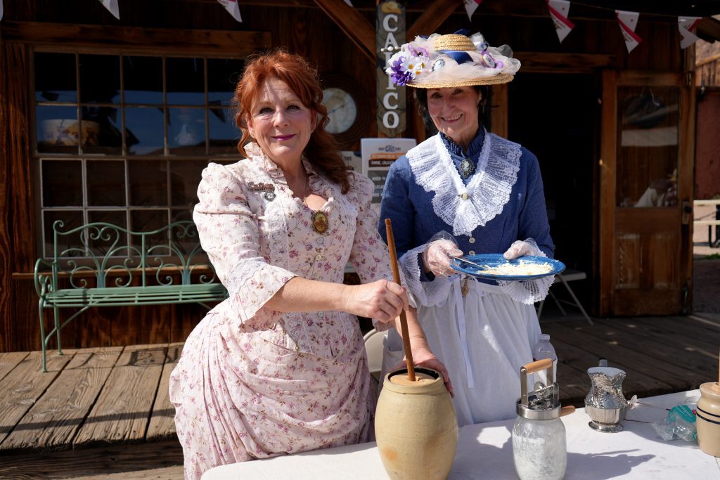Two women in period clothing stand at a table with a butter churn.