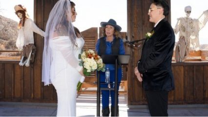 Two people face each other in a wedding dress and suit with a woman in the middle wearing Western attire with Halloween decorations in the background.