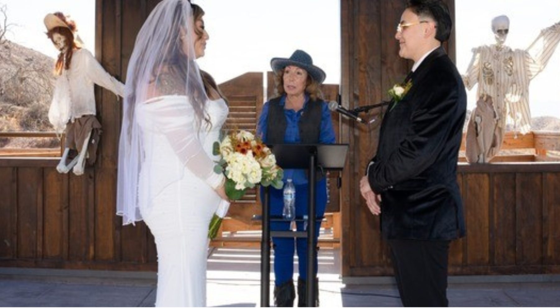 Two people face each other in a wedding dress and suit with a woman in the middle wearing Western attire with Halloween decorations in the background.