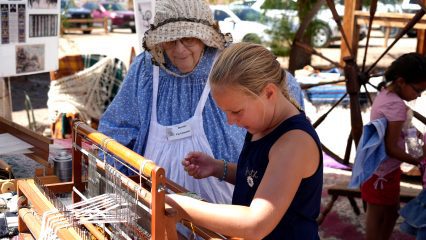 A young girl learns how to weave yarn on a traditional wooden loom during a living-history demonstration at Calico Days. A woman dressed in period-style clothing guides her as another child participates nearby.