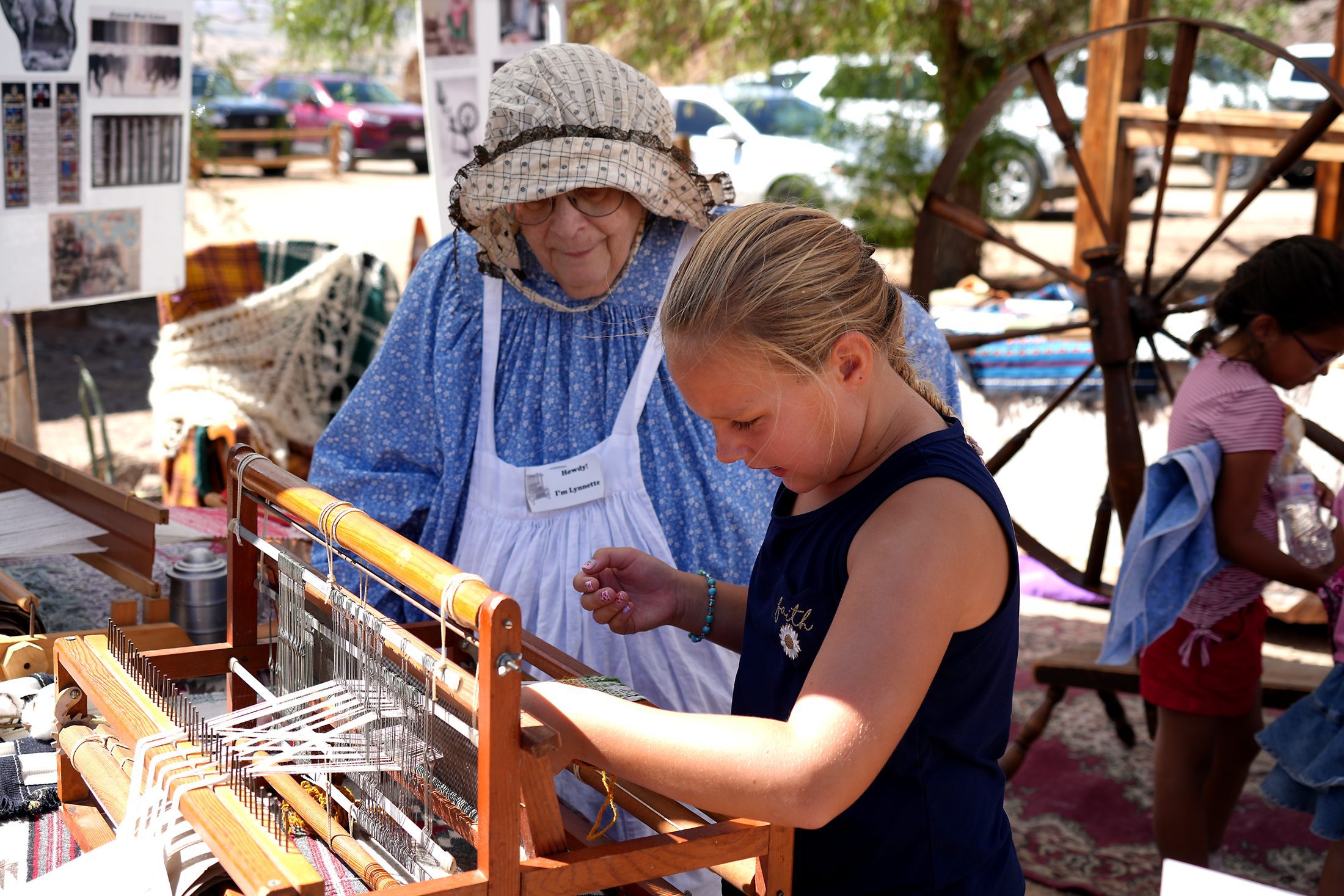A young girl learns how to weave yarn on a traditional wooden loom during a living-history demonstration at Calico Days. A woman dressed in period-style clothing guides her as another child participates nearby.